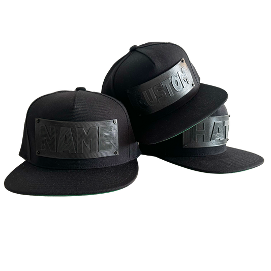 3 snapback baseball caps in black with black 3D printed logo panels. One says "CUSTOM",one says NAME", and the third says "HAT".