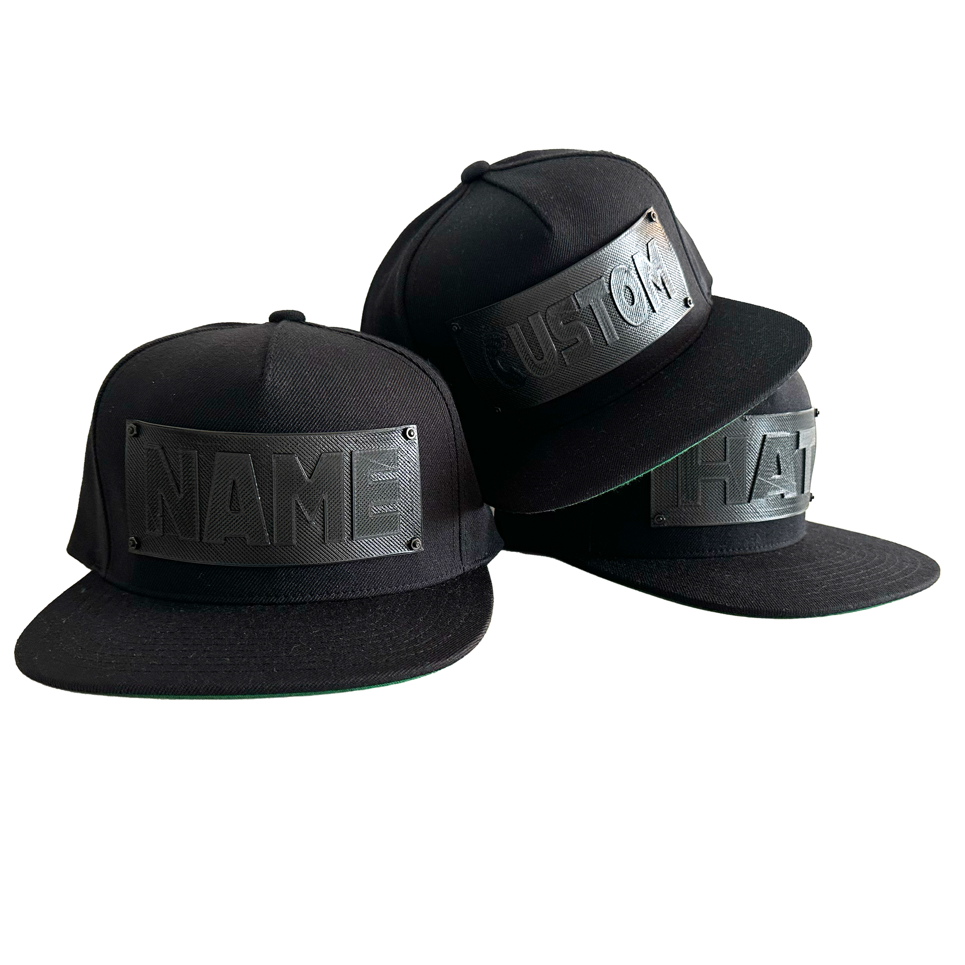 3 snapback baseball caps in black with black 3D printed logo panels. One says "CUSTOM",one says NAME", and the third says "HAT". 