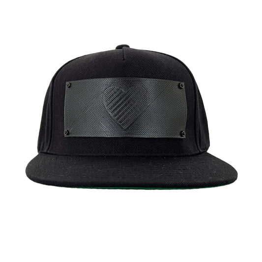 A black snapback baseball hat with a  rectangular 3D printed black logo panel with a heart symbol on it.