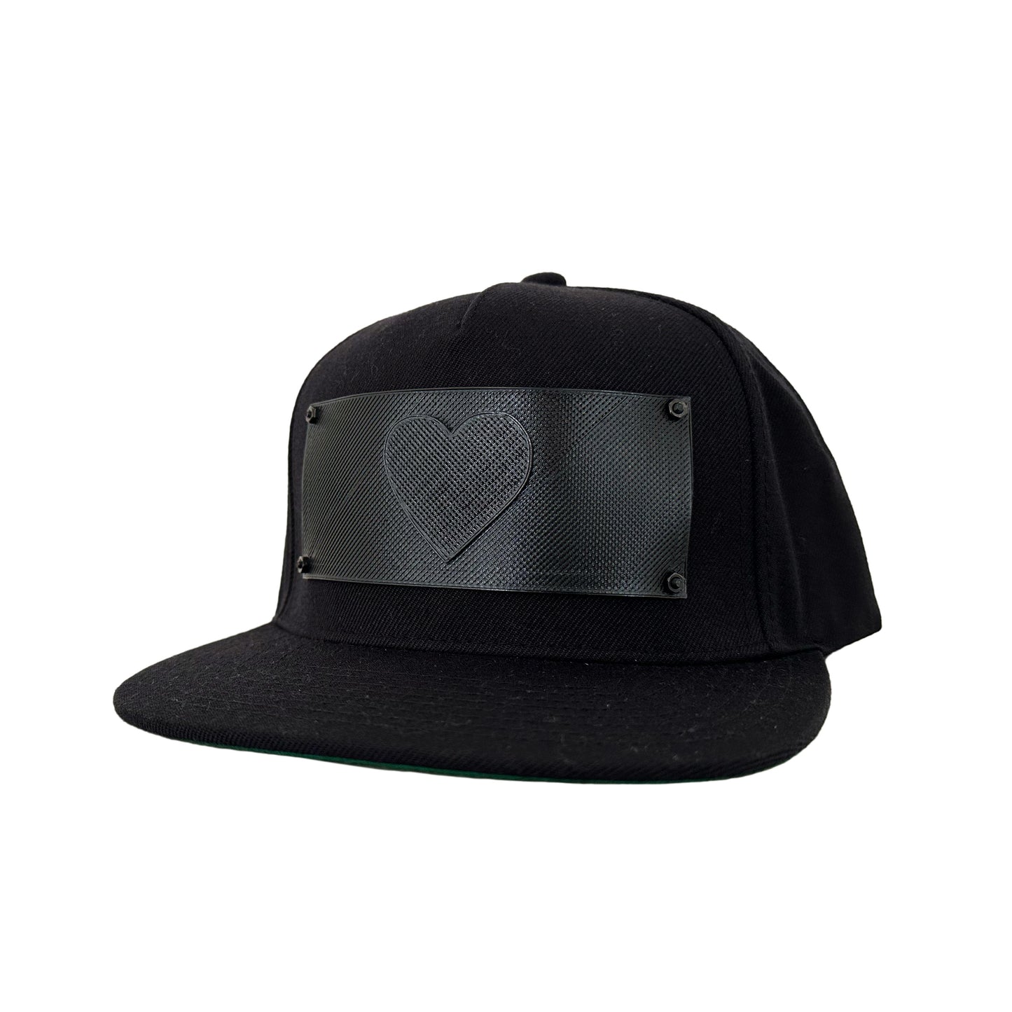 A black snapback baseball hat with a rectangular 3D printed black logo panel with a heart symbol on it.