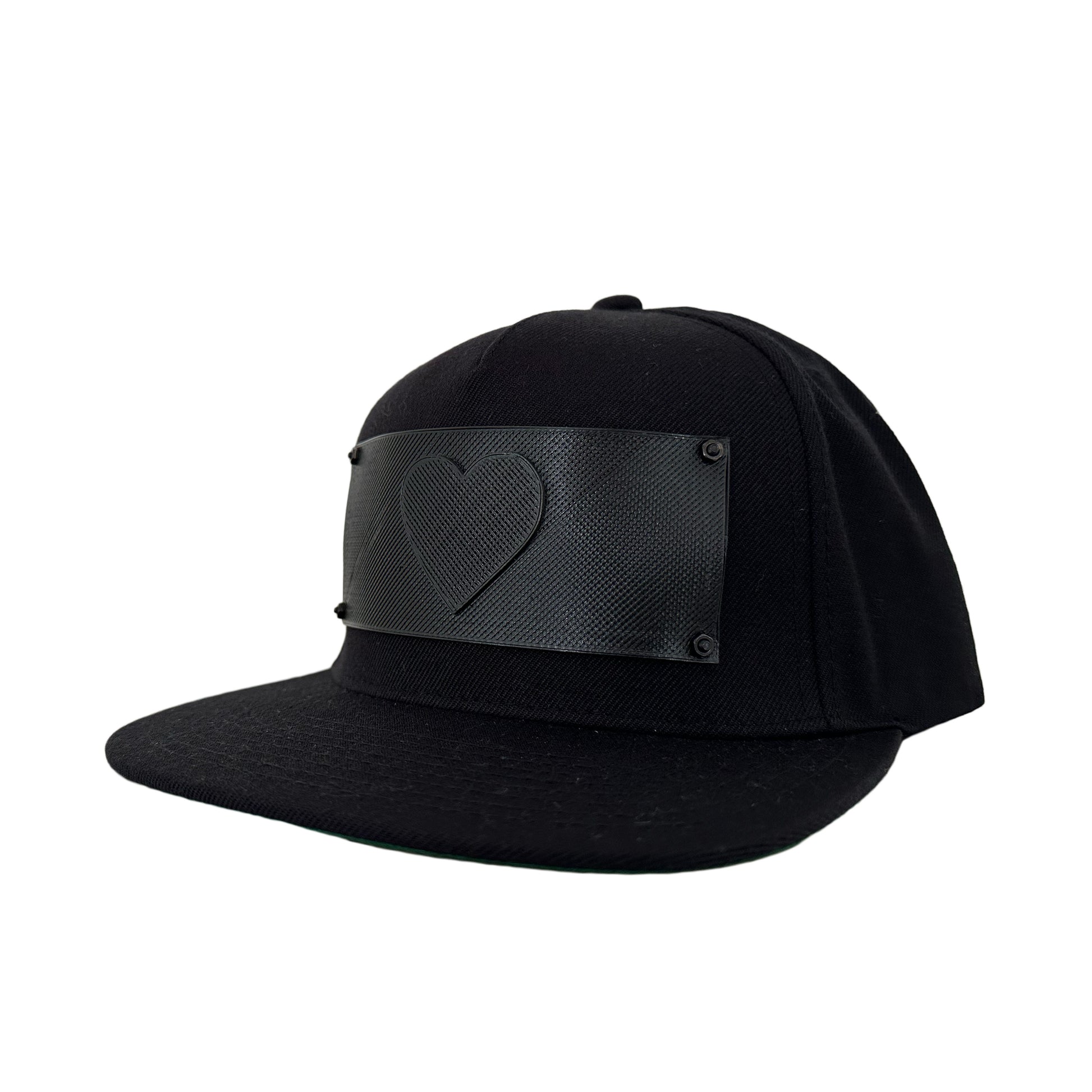 A black snapback baseball hat with a rectangular 3D printed black logo panel with a heart symbol on it.